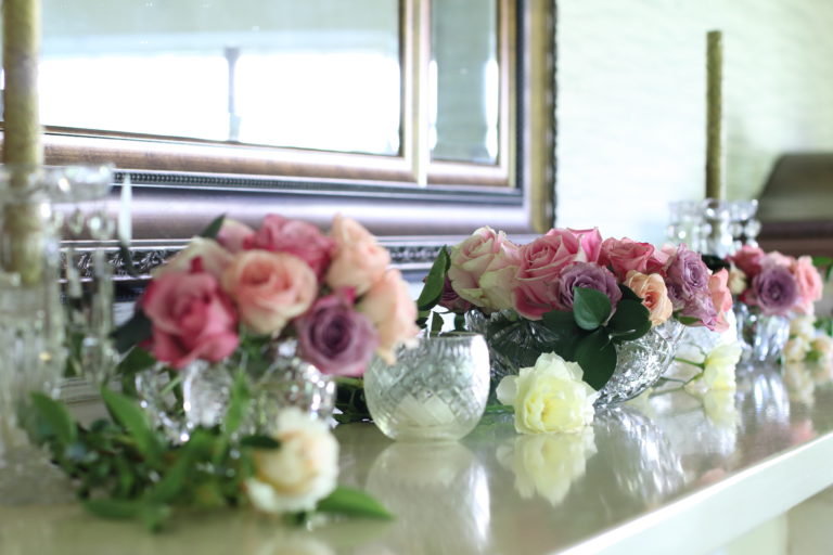 Angle view of the mantel with pink roses in crystal vases, cream & soft yellow roses lying on the mantel, & votive candle holders.