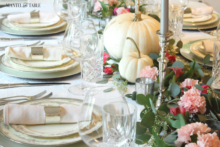 Close shot of china with pink flowers on silver chargers. Blush linen napkins rolled in sleek silver rings. White pumpkin & pink flower garland w/silver candles.