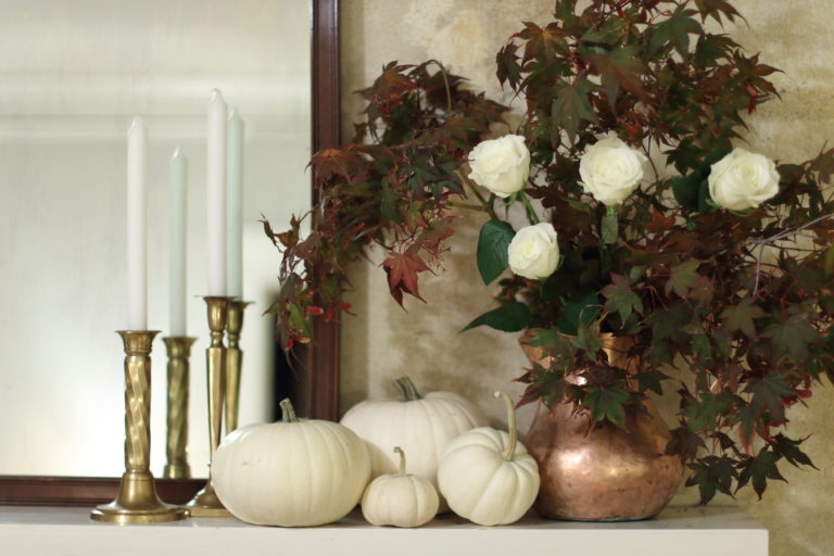 White pumpkins on the mantel. Copper pitcher with fall leaves & white roses on the right, brass candle sticks & white candles on the left in front of a dark wood mirror.