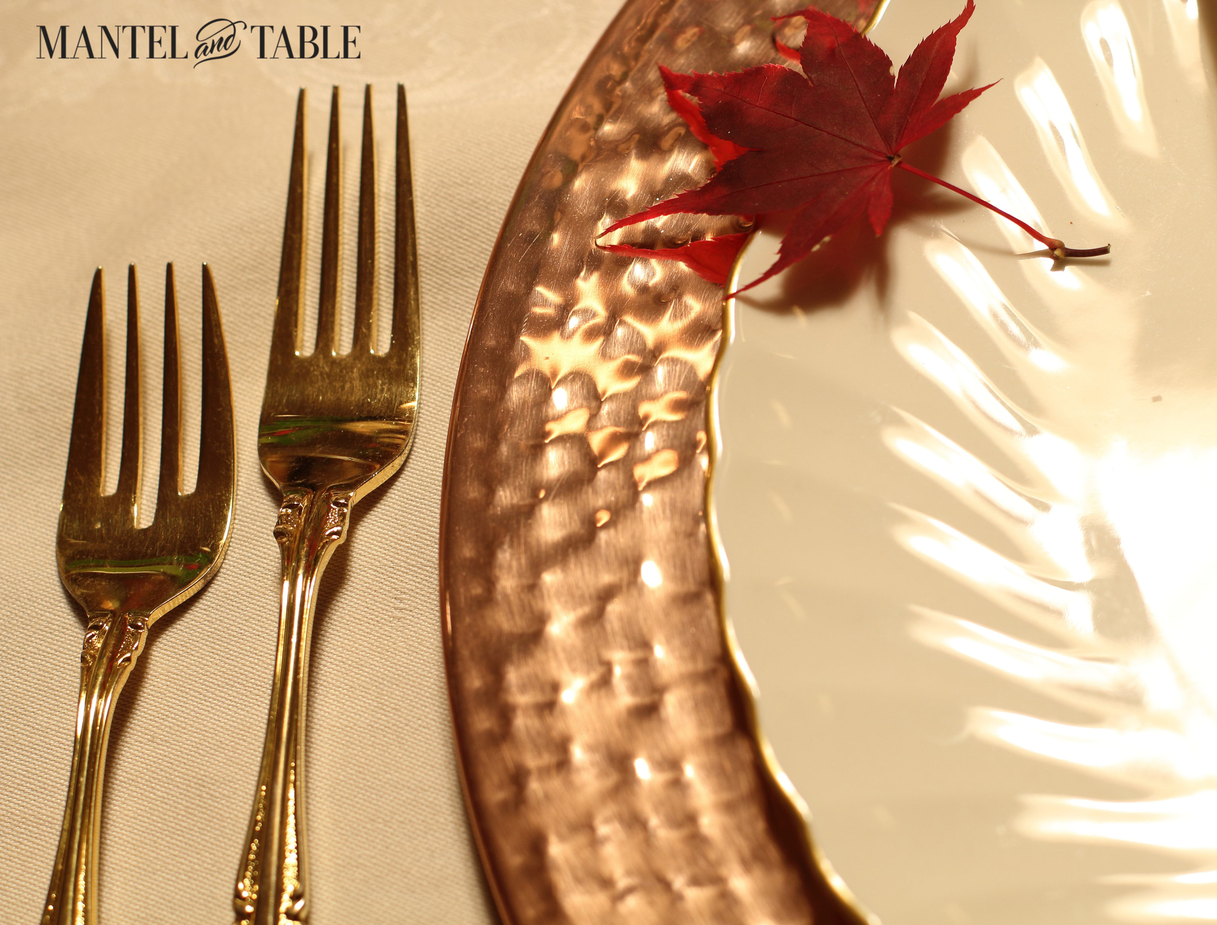 Copper charger, white scalloped plate with gold rim, gold forks to the left and a small red maple leaf on the plate