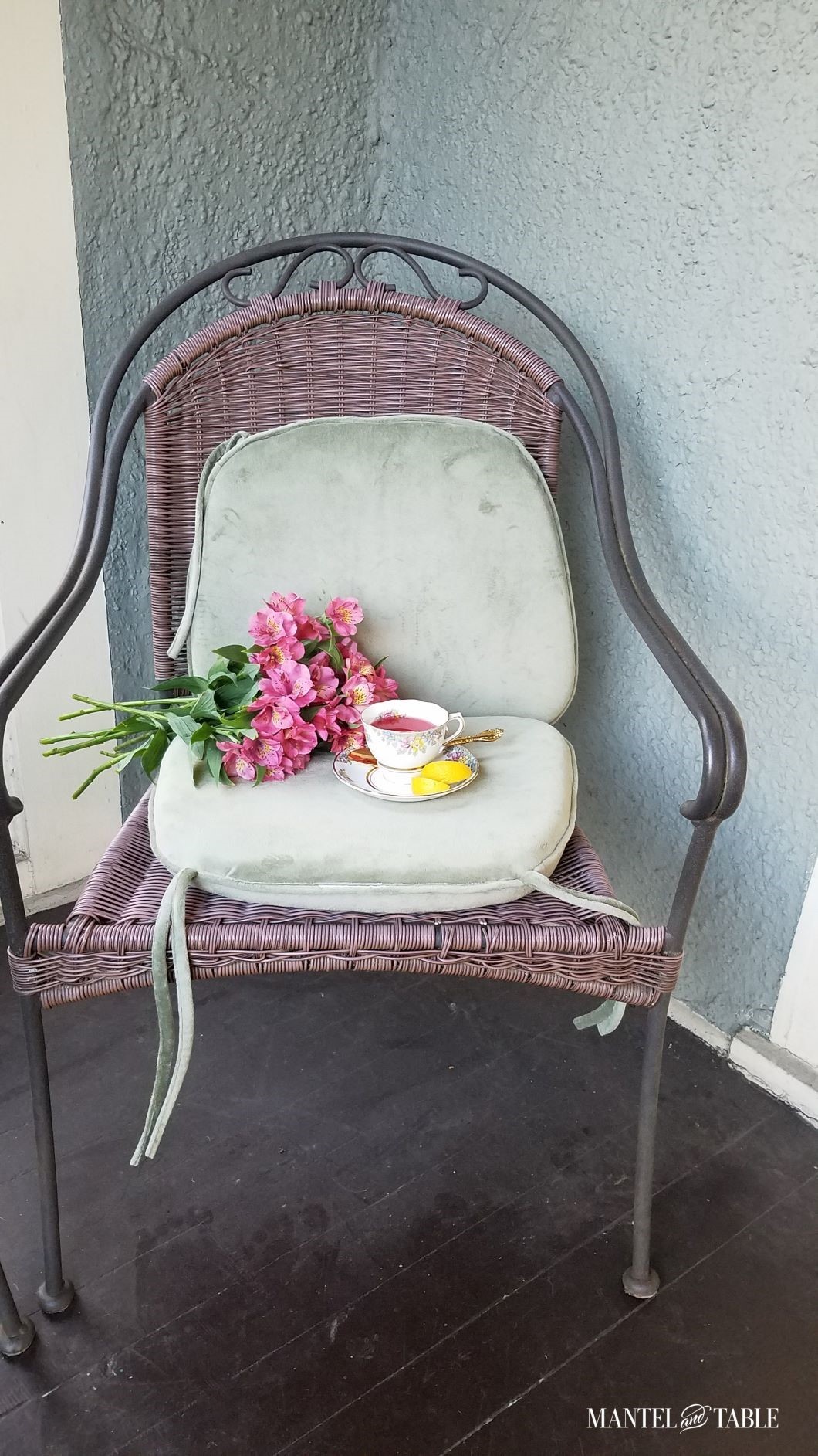 Distance view of patio chair on porch with green seat cushions, bouquet of pink flowers, and cup & saucer with red tea and lemon slices